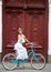 Beautiful female in white dress posing on blue vintage bike in front of beautiful old red doors