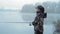 Beautiful fisherwoman girl in camouflage costume throws fishing rod into the river in foggy autumn morning