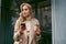 Beautiful flirty casual blond girl in stylish trench coat with coffee to go and cellphone dreamily looking away outdoor
