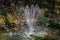 Beautiful fountain in autumn garden pond against background of shady fall garden. Freshness of water jets