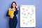 Beautiful and full of energy young confident woman holding yellow paper folder in hands, standing near flipchart with multi