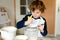 Beautiful funny blond little kid boy baking chocolate cake and tasting dough in domestic kitchen