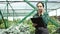 Beautiful gardener girl in apron holding clipboard and talking on phone stands among green plants in greenhouse. Farmer