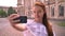 Beautiful ginger young woman with bare shoulders taking selfie with her phone and smiling, standing on street with
