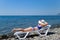 A beautiful girl in a blue bikini and a white hat is sunbathing on the beach, lying on a white chaise longue. The blonde