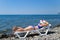 A beautiful girl in a blue bikini and a white hat is sunbathing on the beach, lying on a white chaise longue. The blonde