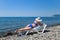 A beautiful girl in a blue bikini and a white hat is sunbathing on the beach, lying on a white chaise longue. The blonde