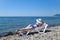 A beautiful girl in a blue bikini and a white hat is sunbathing on the beach, lying on a white chaise longue. The blonde