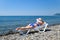 A beautiful girl in a blue bikini and a white hat is sunbathing on the beach, lying on a white chaise longue. The blonde