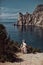 A beautiful girl, a bride, in a white dress sits a tree, on a cliff against the background of the ocean and mountains.