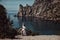 A beautiful girl, a bride, in a white dress sits a tree, on a cliff against the background of the ocean and mountains.