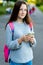 Beautiful girl brunette schoolgirl. Summer in nature. Behind back is pink backpack. In hands of phone with headphones