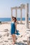 A beautiful girl in a dress, hat and sunglasses poses with a view of the ruins of the ancient Roman temple of Apolon. Side, Turkey