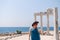 A beautiful girl in a dress, hat and sunglasses poses with a view of the ruins of the ancient Roman temple of Apolon. Side, Turkey