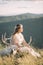 Beautiful girl in a dress sitting with a deer skull with horns on the top of the mountain, eyes closed, decoration on your head