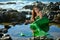 A beautiful girl in a green mermaid costume lies in the water among the stones on the beach and looks at the camera