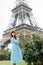 Beautiful girl in a hat and elegant blue dress posing near bush of roses outdoor, walking in Paris. Eiffel tower on the