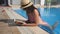 Beautiful girl in hat and sunglasses reading book at pool. Young woman relaxing at warm sunny day during vacation