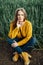 Beautiful girl with jeans, a yellow sweater and boots sits near an early wheat field