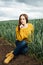 Beautiful girl with jeans, a yellow sweater and boots sits near an early wheat field