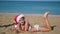 A beautiful girl lies on the sand by the sea and waves her legs. A child wearing a santa claus hat is celebrating Christmas