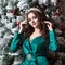 A beautiful girl with long hair in a green dress with the white diadem on her head near the Christmas tree . A square picture.