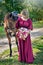 A beautiful girl in a long red dress is standing next to a brown horse.