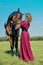 Beautiful girl in a long red dress stands next to the brown horse.