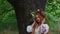 Beautiful girl with long red hair with flower crown in white and red embroidered ethnic suit stands by tree
