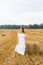 Beautiful girl in a long white dress and barefoot standing at setga of straw on a yellow field