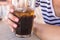 Beautiful girl with a pink manicure holds and drinking a large glass with soda with pieces of ice and a straw on a sunny hot day.