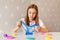 A beautiful girl is playing with colored slime at a table in the room. The child looks at the blue slime and stretches it to