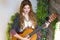 A beautiful girl with red curly hair stands and holds a seven-string guitar in her hands.