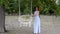 A beautiful girl with red hair in a blue dress poses for the camera near a white swing set in an empty city Park