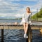 A beautiful girl sits alone on a river pier under a clear sun and looks at the water. The wind develops her blond hair curls.