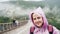 Beautiful girl smiles in a pink sweatshirt in a hood, a backpack and a raincoat in drops on a bridge in Montenegro