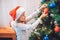 Beautiful girl standing at Christmas tree and dressing her with colorful toys. She holds red one with both hands. Girl