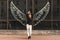 Beautiful girl stands with his hands raised against the background of a dark wall where the wings are drawn, looking in camera and