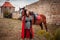 A beautiful girl stands next to a horse against the background of a medieval fortress. Fantasy costume, chain mail, cloak. A woman