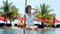 Beautiful girl with tropical cocktail sitting at outdoor swimming pool with sea, palm trees and sun lounges on