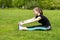 Beautiful girl warms up muscles before training. Sitting on the grass in the park stretches arms to legs.