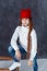 A beautiful girl wearing a red hat with kitten ears. A girl with very ling hair in a studio sitting on the wooden cube