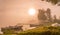 Beautiful golden Sun glows through fog at lake during early sunrise, boats and long wooden bridge, trees, sky reflection in calm