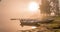 Beautiful golden Sun glows through fog at lake during early sunrise, boats and long wooden bridge, trees, sky reflection in calm