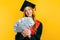 Beautiful graduate girl, in a black gown, and a black master`s hat, with dollars in her hands, marks the graduation, on a yellow