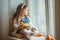 Beautiful happy child girl is sitting on window sill with her friend rabbit and holding basket with painted eggs, Easter