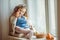 Beautiful happy child girl is sitting on window sill with her friend rabbit and holding basket with painted eggs, Easter