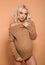 Beautiful happy pregnant woman expecting maternity hugging her belly. Blonde in knit sweater isolated on studio beige background.