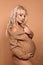 Beautiful happy pregnant woman expecting maternity hugging her belly. Blonde in knit sweater isolated on studio beige background.