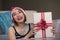 Beautiful and happy woman in Santa hat holding Christmas present box with red ribbon smiling excited at home living room receiving